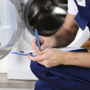 ApplianceSolutions-appliance-maintenance-041620251 main crouching down near a washing machine with a pen and clipboard - Appliance Solutions appliance maintenance